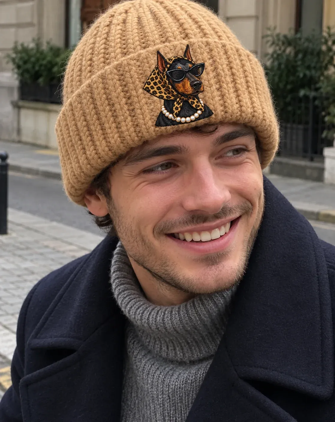 A smiling man in a navy pea coat wearing the camel Noodle beanie, a doberman in a leopard headscarf on the cuff